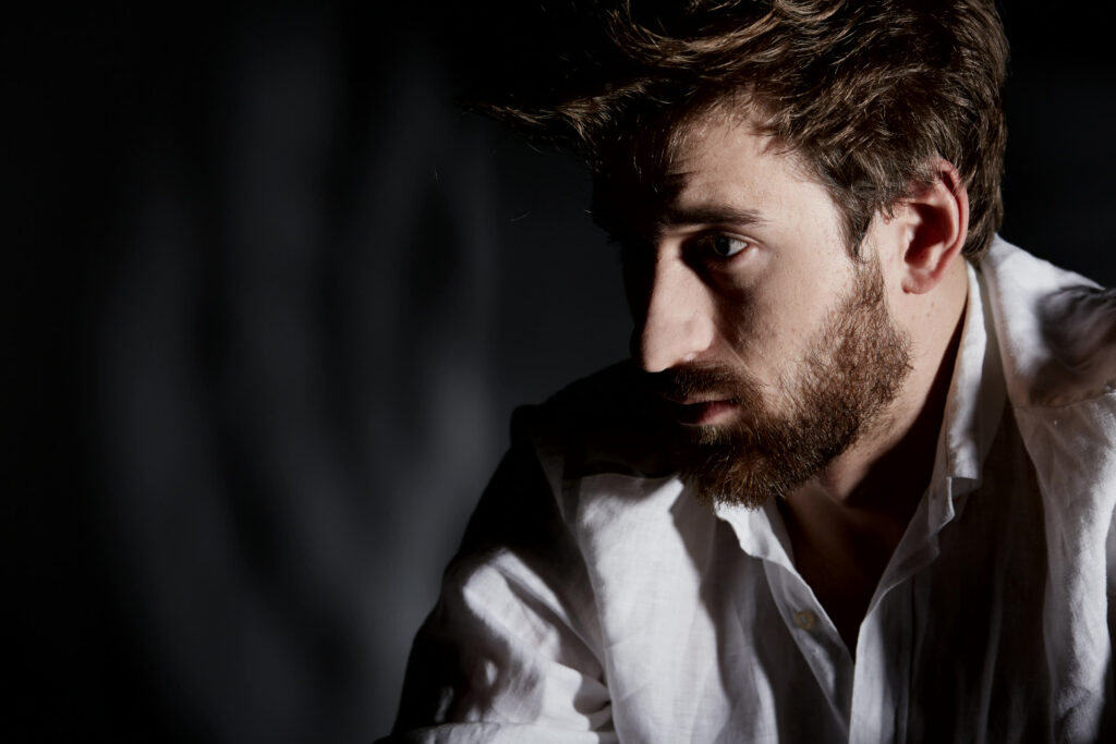 Man with a short, well-kept beard in low, moody side light against a dark background