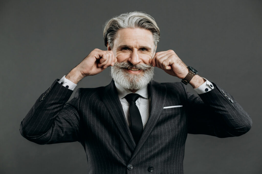 Man in a suit with a grey beard twisting the ends of his styled moustache against a dark grey backdrop